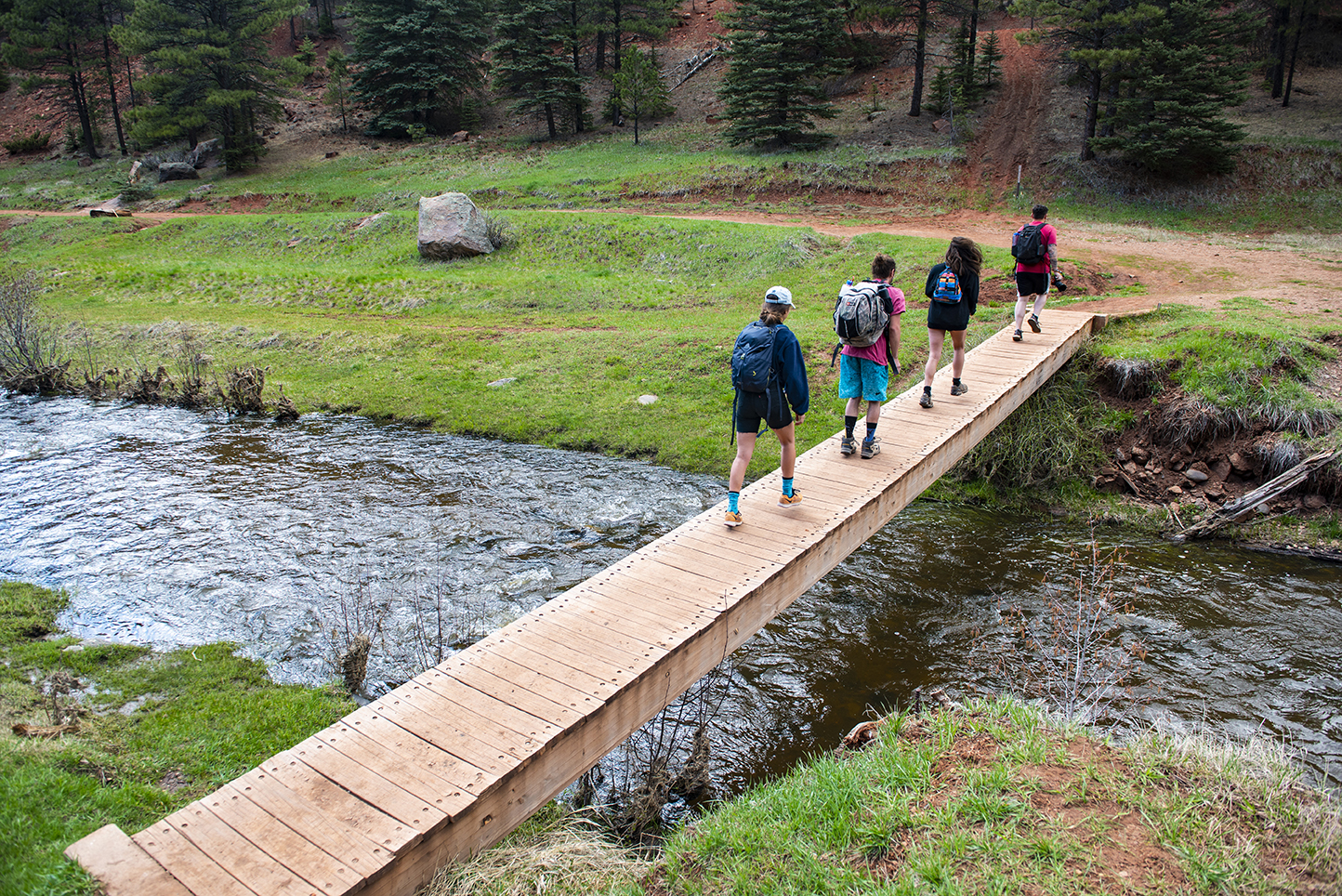 photo: Men and women wearing backpacks cross a river on a wooden bridge